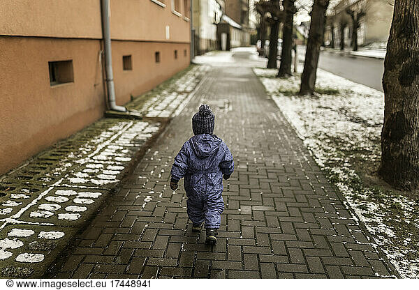 Little boy in blue onesie and winter hat walking on pavement