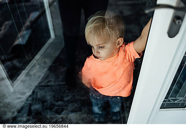 Little boy in bright shirt looks longingly out smudged door at t