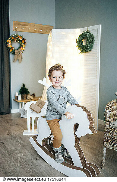 Little boy sitting on wooden horse in Christmas decorated room.