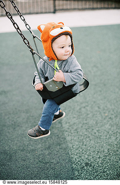 Little boy with fox hat swinging at the playground.