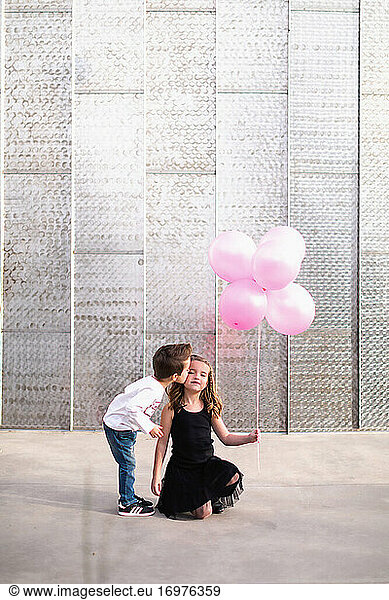 Little brother kissing sister holding pink balloons on the cheek.