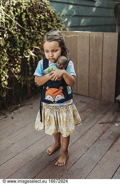 Little girl carrying babydoll in baby carrier outside in yard