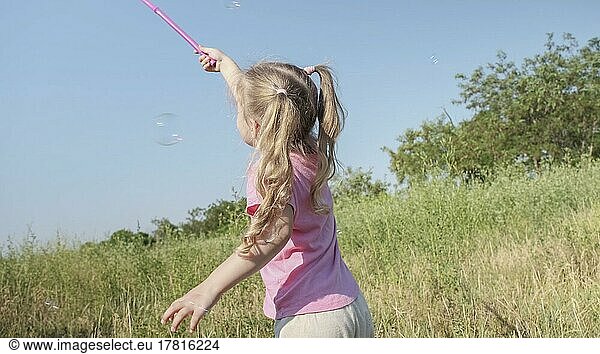 Little girl catches soap bubbles in butterfly net while playing in tall grass in park. Cute little girl smiles and catches on soap bubbles in aerial insect net in meadow on sun day. Odessa  Ukraine  Europe