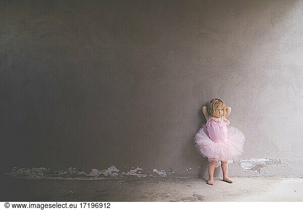 Little girl dressed in a too big ballerina outfit leaning against wall
