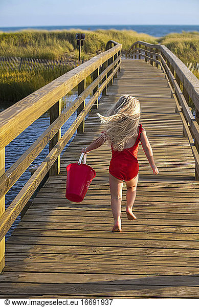 Little Girl From Behind Running On Bridge to Beach with Red Bucket