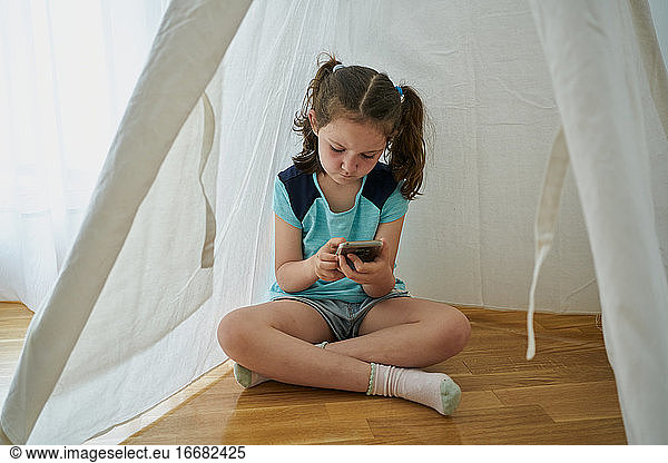 Little girl looking at her smartphone inside a white teepee tent inside her house. Technology concept