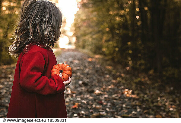 Little girl outside in a fall forest holds a tiny pumpkin in her hand