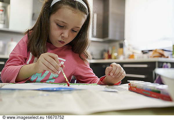 Little girl painting a drawing with watercolors inside her house with a kitchen in the background. Creative concept