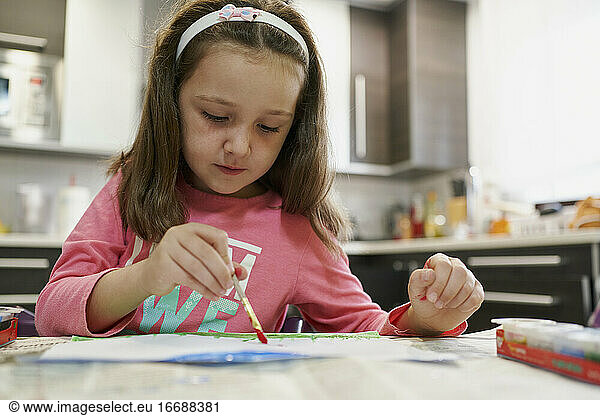 Little girl painting a drawing with watercolors inside her house with a kitchen in the background. Creative concept