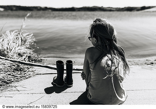 Little Girl Sits Beside Lake With Boots Removed