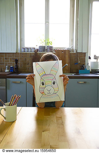 Little girl sitting at kitchen table  holding drawing of Easter bunny in front of face