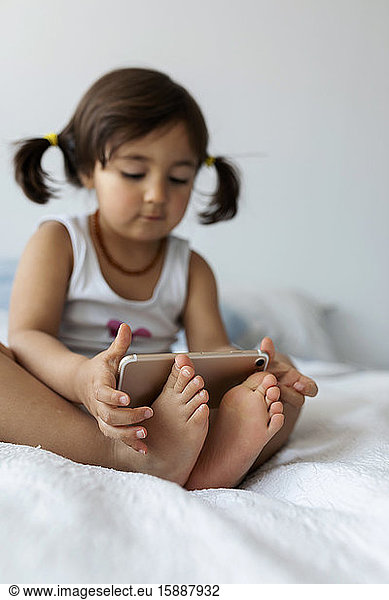 Little girl sitting on bed holding smartphone with feet  close-up