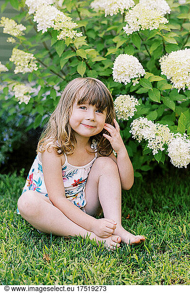 Little girl sitting on lawn in front of hydrangea bush