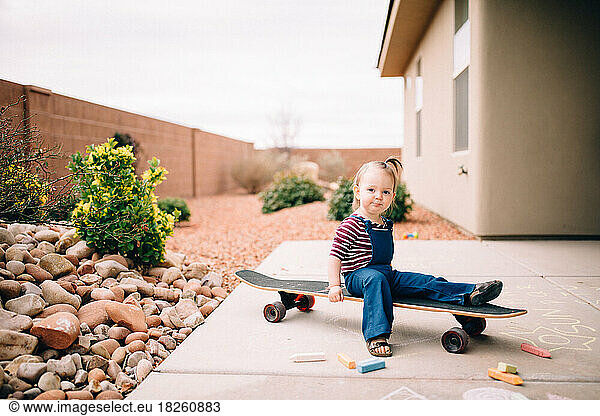 Little girl sitting on longboard and coloring with chalk outside
