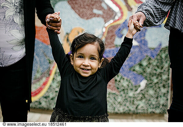Little girl smiling & holding hands in front of color mural