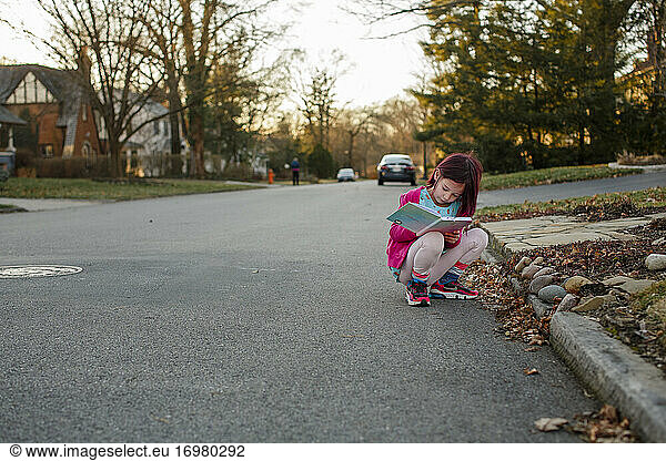 Little girl squats down in street to write notes in journal at sunset