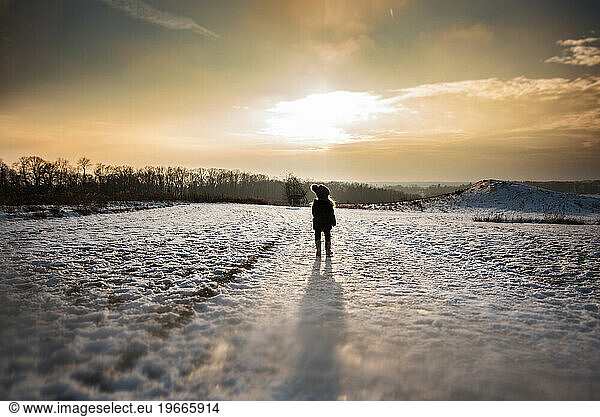 Little girl standing alone in snowy field at sunset