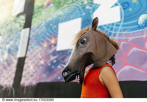 Little girl with a horse's head and a red dress next to a graffiti wall