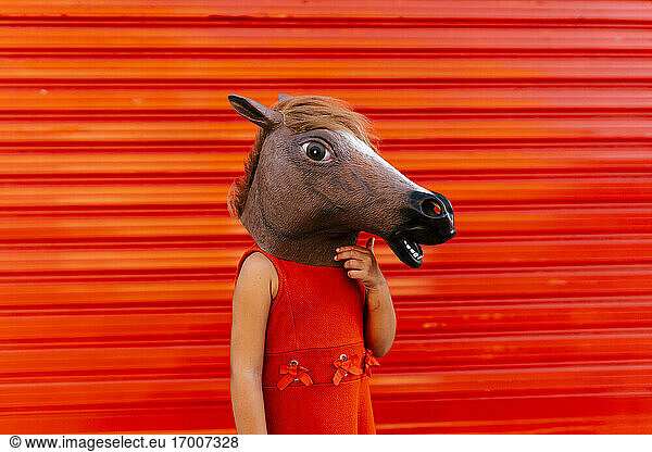 Little girl with a horse's head and a red dress standing in front of red roller shutter