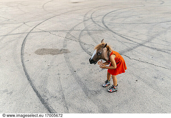Little girl with a horse's head and a red dress  standing on asphalt with tire tracks
