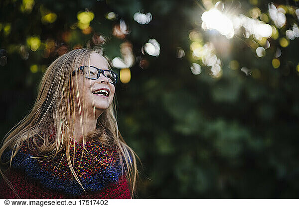 Little girl with long hair and glasses laughs in front of fall foliage