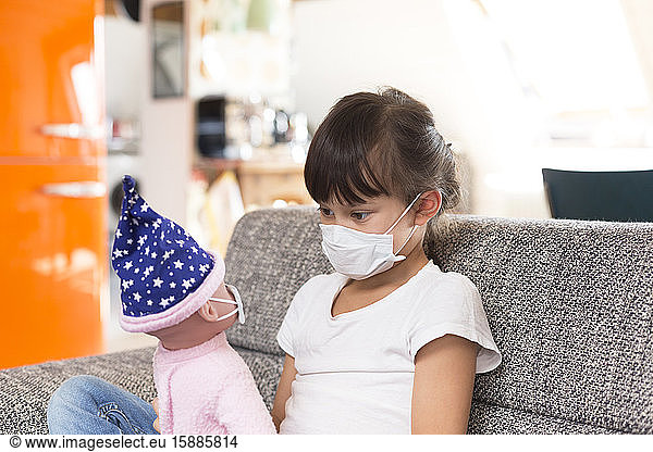 Little girl with protective mask sitting on couch at home playing with doll