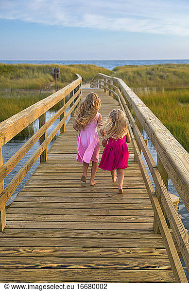 Little Girls From Behind Running On Bridge to Beach in Pink Dresses