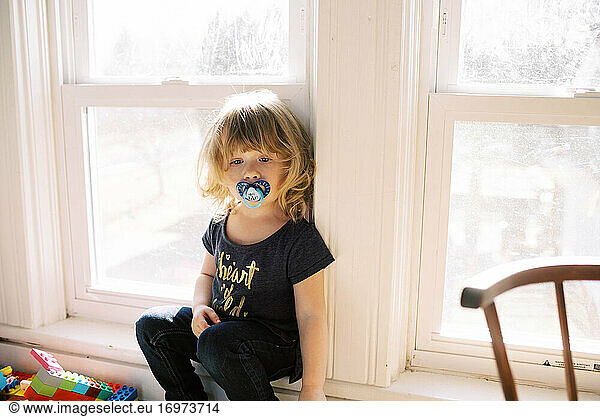 Little tired toddler girl with pacifier sitting in play room by window