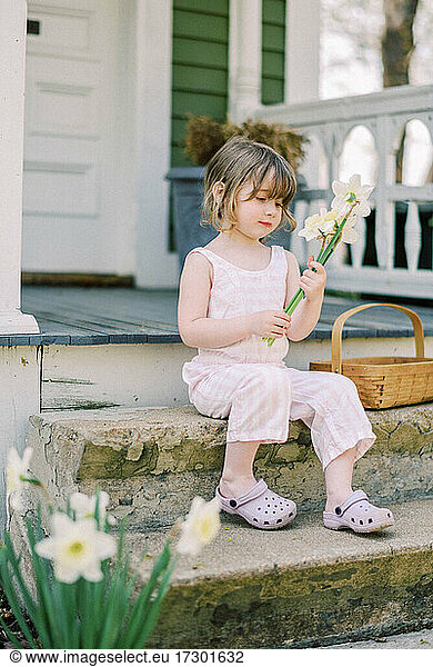 Little toddler girl holding up a bunch of daffodils on porch
