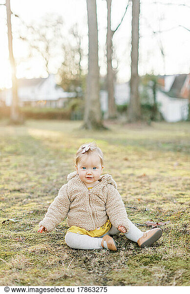 Little toddler girl in bow and jacket sits in a field during sunset