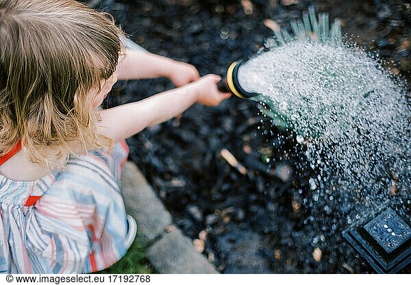 Little toddler girl in summer dress watering blooming garden in spring