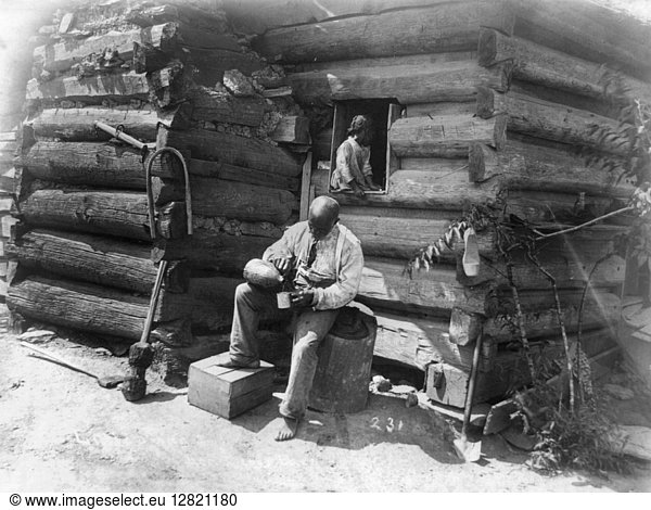 LOG CABIN  c1895. An African American man seated on a tree stump  outside a log cabin in rural America while pouring a drink into cup with a young girl seated in the window above him. Photograph  c1895.