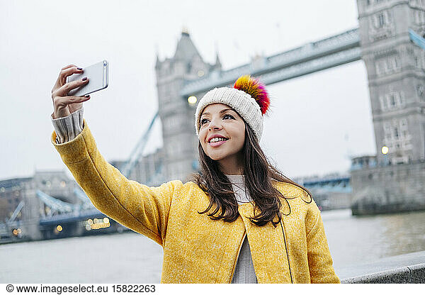 London  United Kingdom  Girl with a wooly hat and yellow coat takes a selfie in front of Tower Bridge