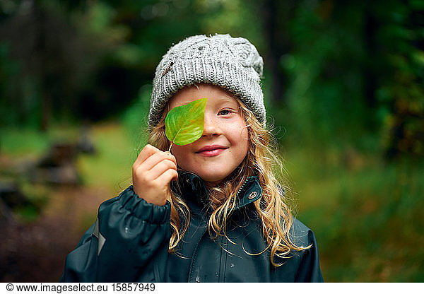 Long haired charming girl in warm hat closing eye with green leaf in forest