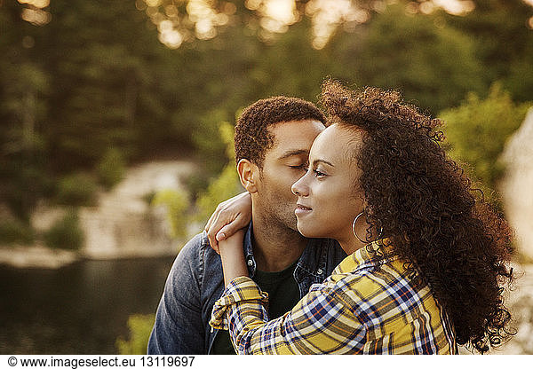 Loving man kissing woman while sitting at lakeshore during sunset
