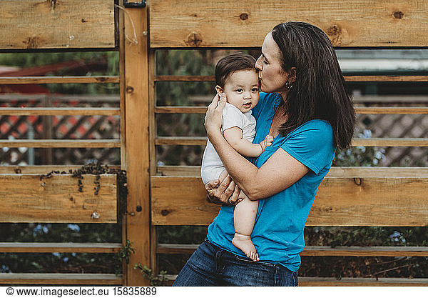 Loving mom kissing forehead of baby girl outdoors near fence