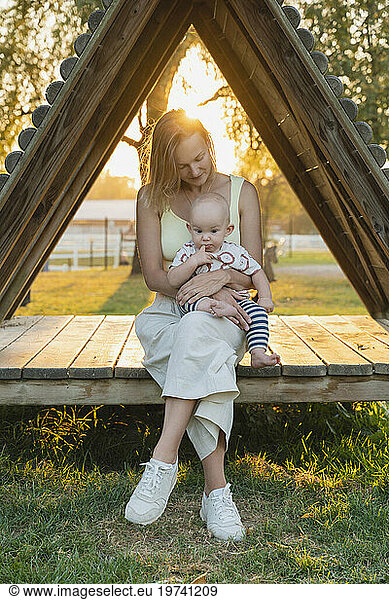 Loving mother sitting with baby boy in triangle wooden seat at sunset