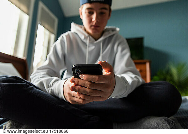 Low angle of teen boy in white sweater on smartphone in bedroom.