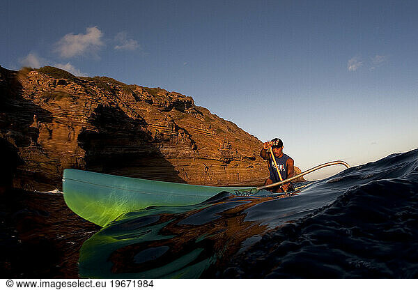 Low angle perspective of a man paddling a one man outrigger canoe in nice light next to blg cliffs.