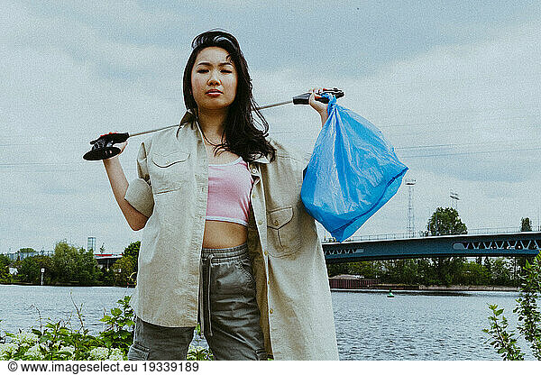 Low angle portrait of confident young woman picking garbage near river