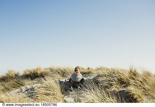 Low angle view of boy sitting on sand dune against clear blue sky
