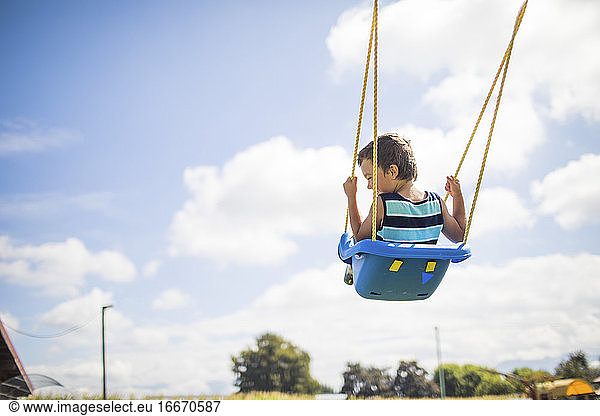 Low angle view of boy swinging outdoors on blue swing at playground.