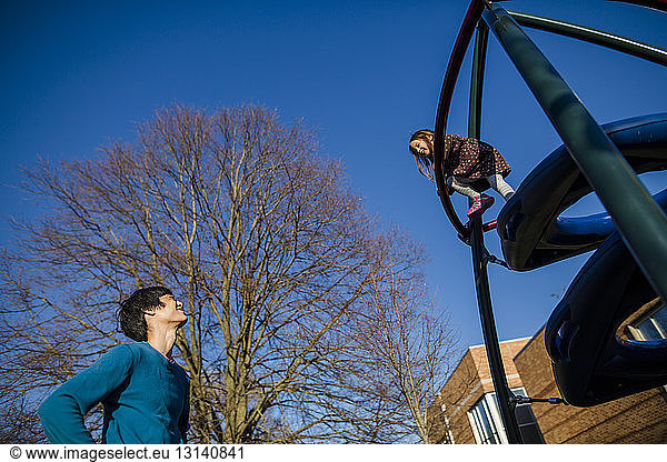 Low angle view of father looking at daughter climbing on outdoor play equipment at playground