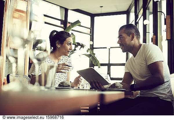 Low angle view of male and female with digital tablet talking while sitting at table in cafe