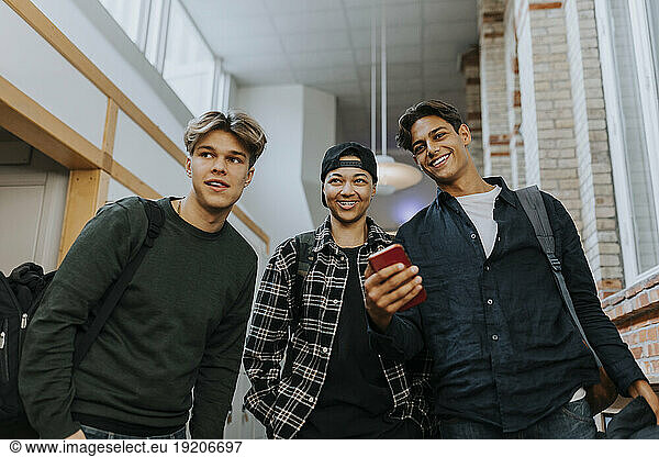 Low angle view of smiling male friends with mobile phone while walking in school corridor