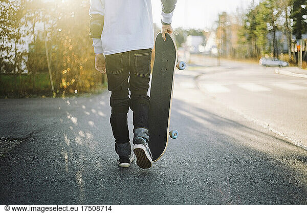 Low section of boy with skateboard walking on road