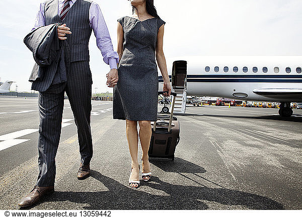 Low section of business couple holding hands while walking on airport runway