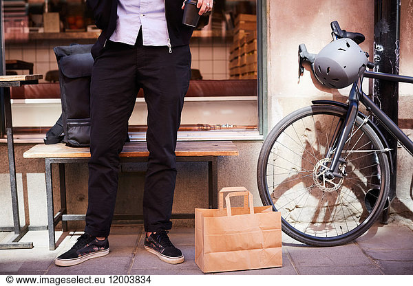 Low section of businessman standing by paper bag and bicycle on sidewalk at city