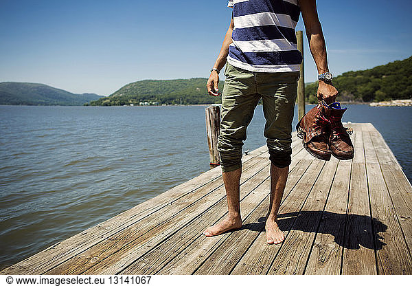 Low section of man holding shoes and standing on jetty over lake