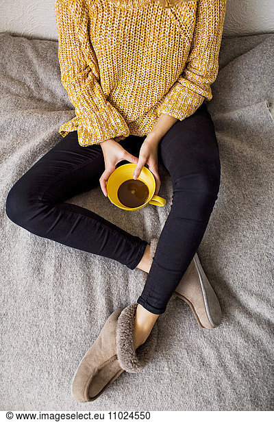 Low section of woman holding coffee cup while resting on bed